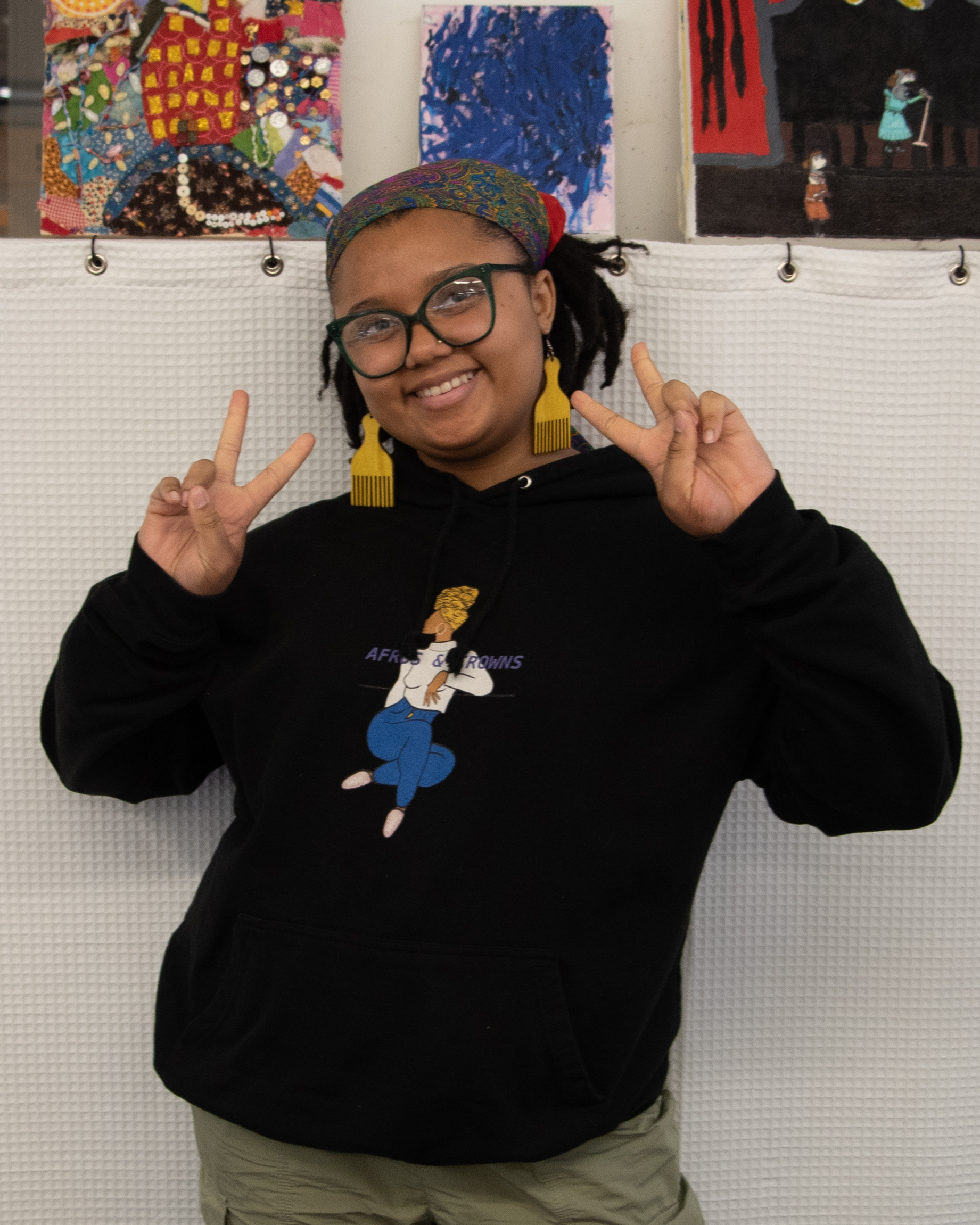 photo of Ketsia Leroy, a young woman with brown skin and dark, textured hair pulled back in a decorative bandana/scarf. She is wearing big glasses, large yellow earrings in the shape of Afro picks and a black, hooded sweatshirt with an image of a digital drawing of a woman and the words Afros and Crowns 